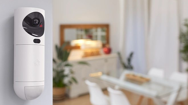 Fotocamera bianca con sensore di movimento fissata a parete, sullo sfondo sala da pranzo luminosa con tavolo in vetro e piante